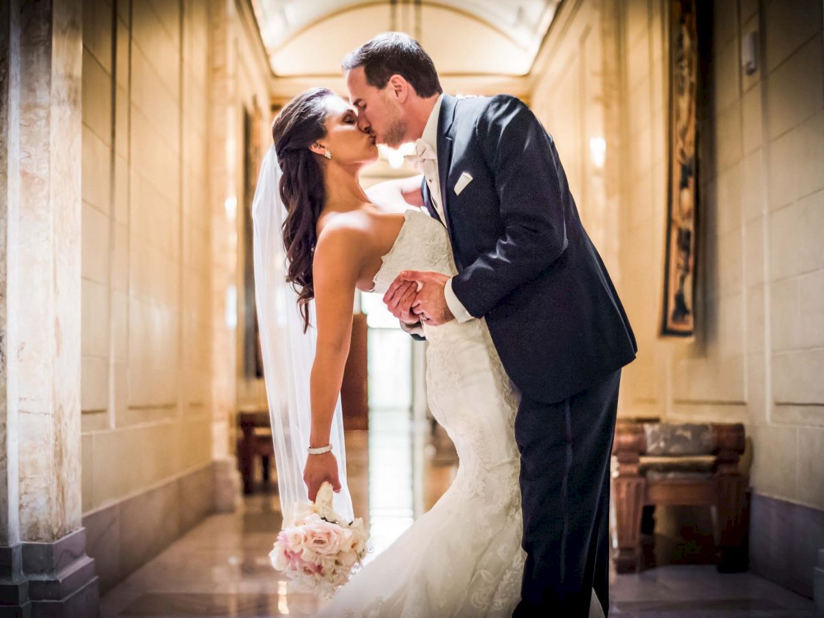 A bride and groom share a romantic kiss in a beautifully lit hallway, with the bride holding a bouquet of flowers.