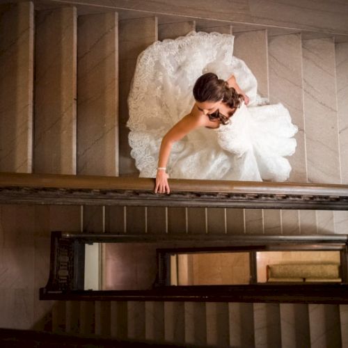 A bride in a white dress is descending a staircase, her hand touching the banister, captured from an overhead perspective.