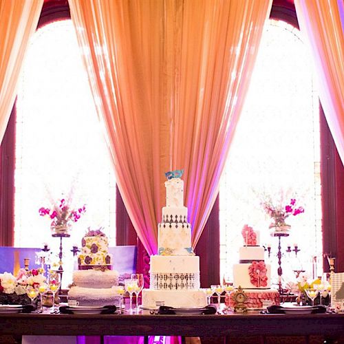 An elegantly decorated table with multiple cakes, floral arrangements, and candles is set against large, draped windows in a lavish setting.