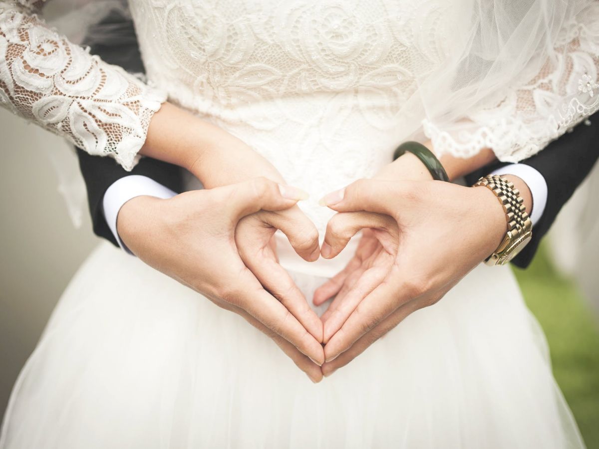 A bride and groom forming a heart shape with their hands, the bride in a lace wedding dress and the groom in a black suit, standing closely together.