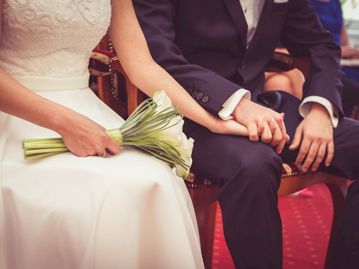 A couple, likely at their wedding, sits closely. The bride holds a bouquet of white flowers while they hold hands on their laps, dressed formally.