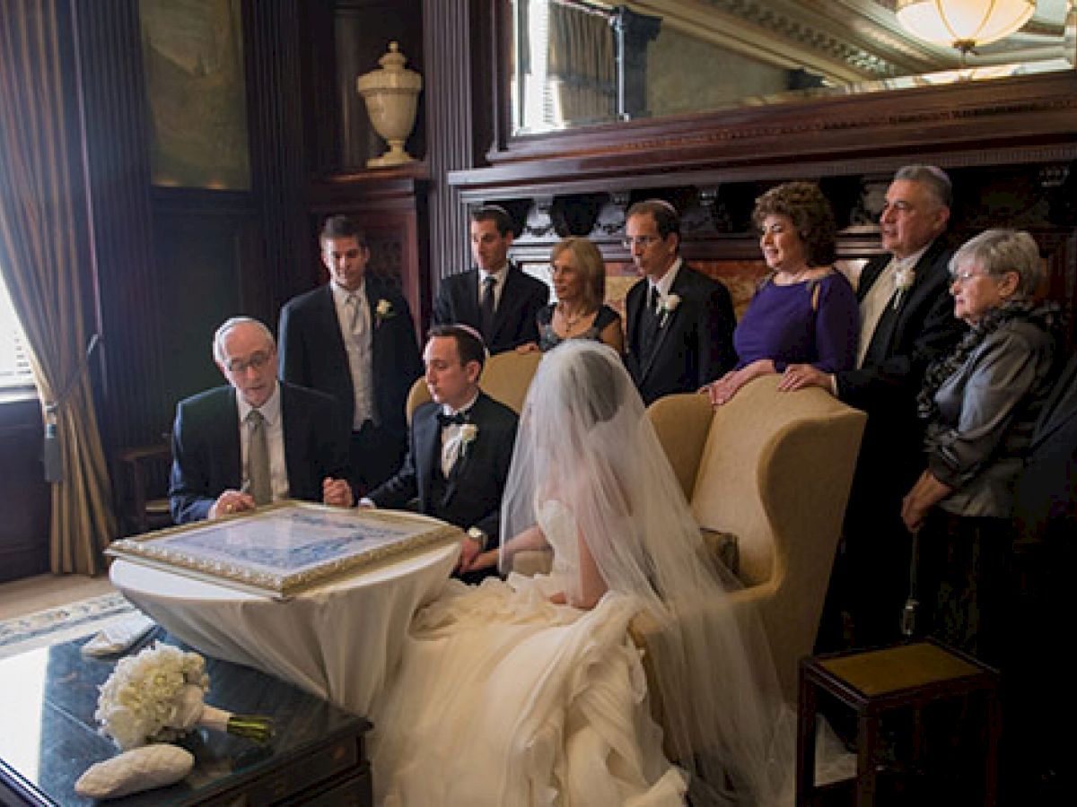 A wedding couple sits with family and friends, signing a document at a formal setting. The bride wears a veil and white dress, surrounded by loved ones.