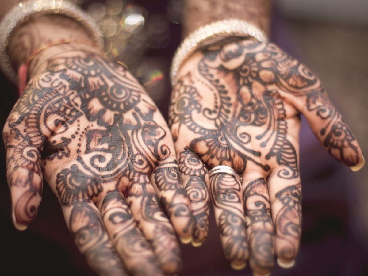 Two hands are decorated with intricate henna designs, featuring traditional patterns and details, and the person is wearing jewelry.