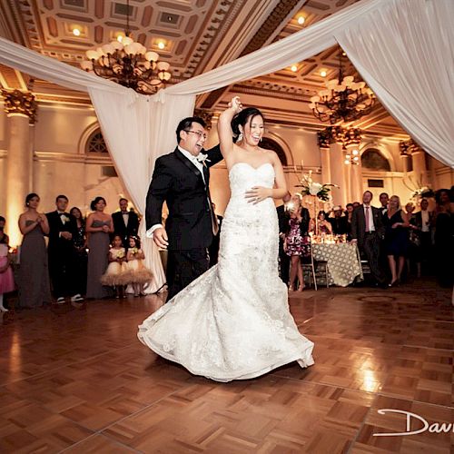 A couple is dancing in a grand ballroom, surrounded by guests and elegant decor, capturing a moment from their special occasion.