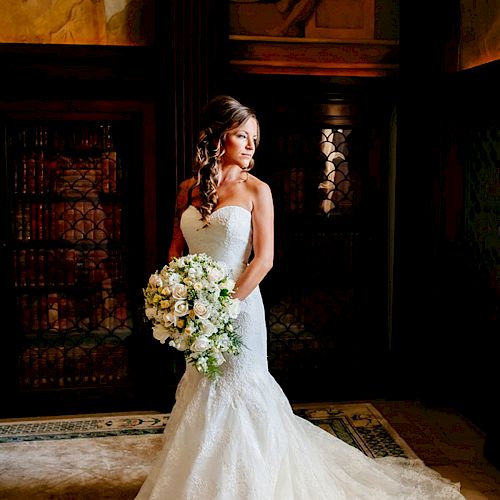 A bride in a white wedding gown holding a bouquet of flowers stands in an ornate room with murals and intricate details.