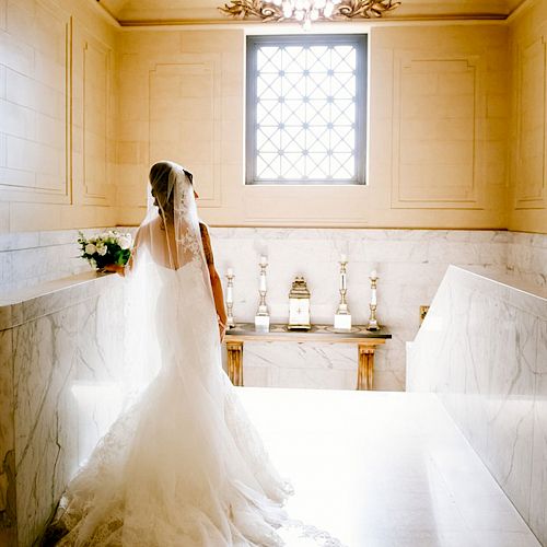 A bride in a white wedding dress and veil stands in a beautifully decorated room with a chandelier and window, facing away from the camera.