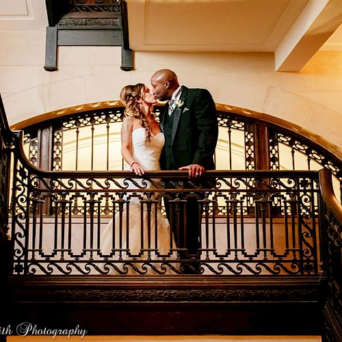 A bride and groom share a kiss on a staircase adorned with ornate railings in a beautifully lit indoor setting.