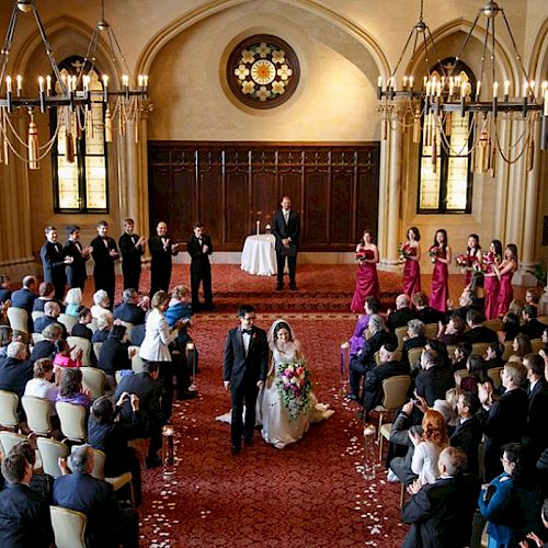 A wedding ceremony in progress inside an elegant room with chandeliers. The couple walks down the aisle as guests and the bridal party look on.