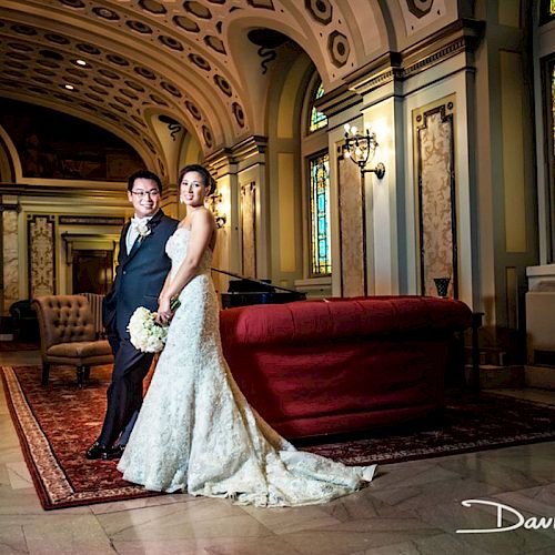 A couple in wedding attire posing indoors in a lavish, ornate room with stained glass windows, high ceilings, and elegant decor.