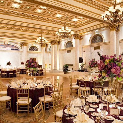 An elegant grand ballroom setup for a formal event with round tables, gold chairs, floral centerpieces, and chandeliers.