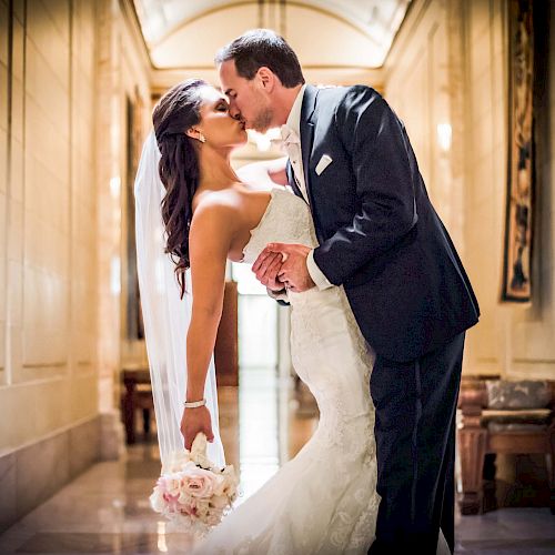 A bride and groom are sharing a kiss in a beautifully lit hallway. The bride holds a bouquet, and the groom is gently dipping her.