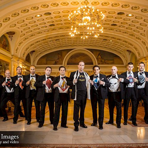 A formal group of men in tuxedos stand in a decorated hall, each holding colorful ties around their necks, under an ornate ceiling and chandelier.