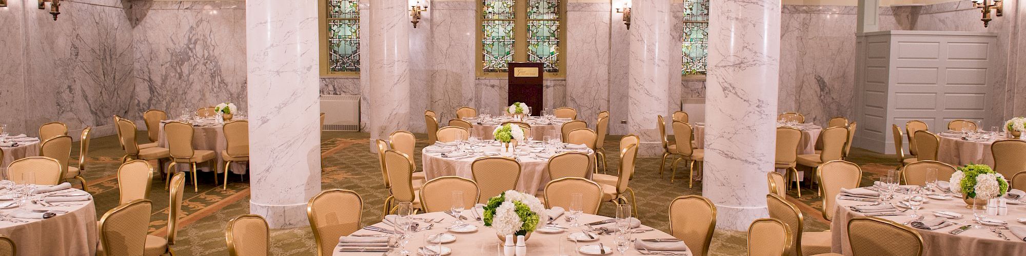 A banquet room with round tables covered in beige tablecloths, set with utensils and centerpiece flowers. Elegant pillars and chandeliers decorate the space.