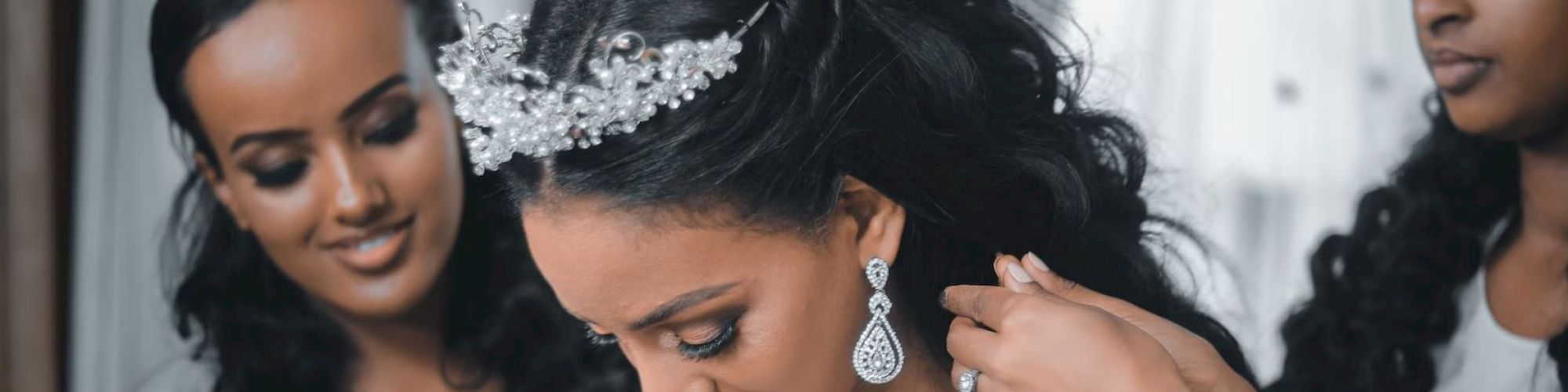 Three women are shown; one wearing a tiara being helped with jewelry by another, while the third smiles and watches.