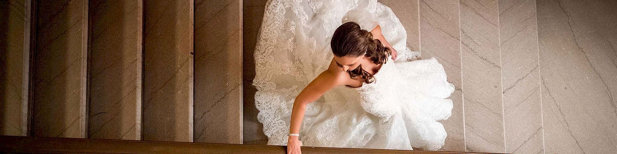 A bride in a white gown descends a staircase, viewed from above, with her hand gliding along the railing and her dress elegantly spread out.