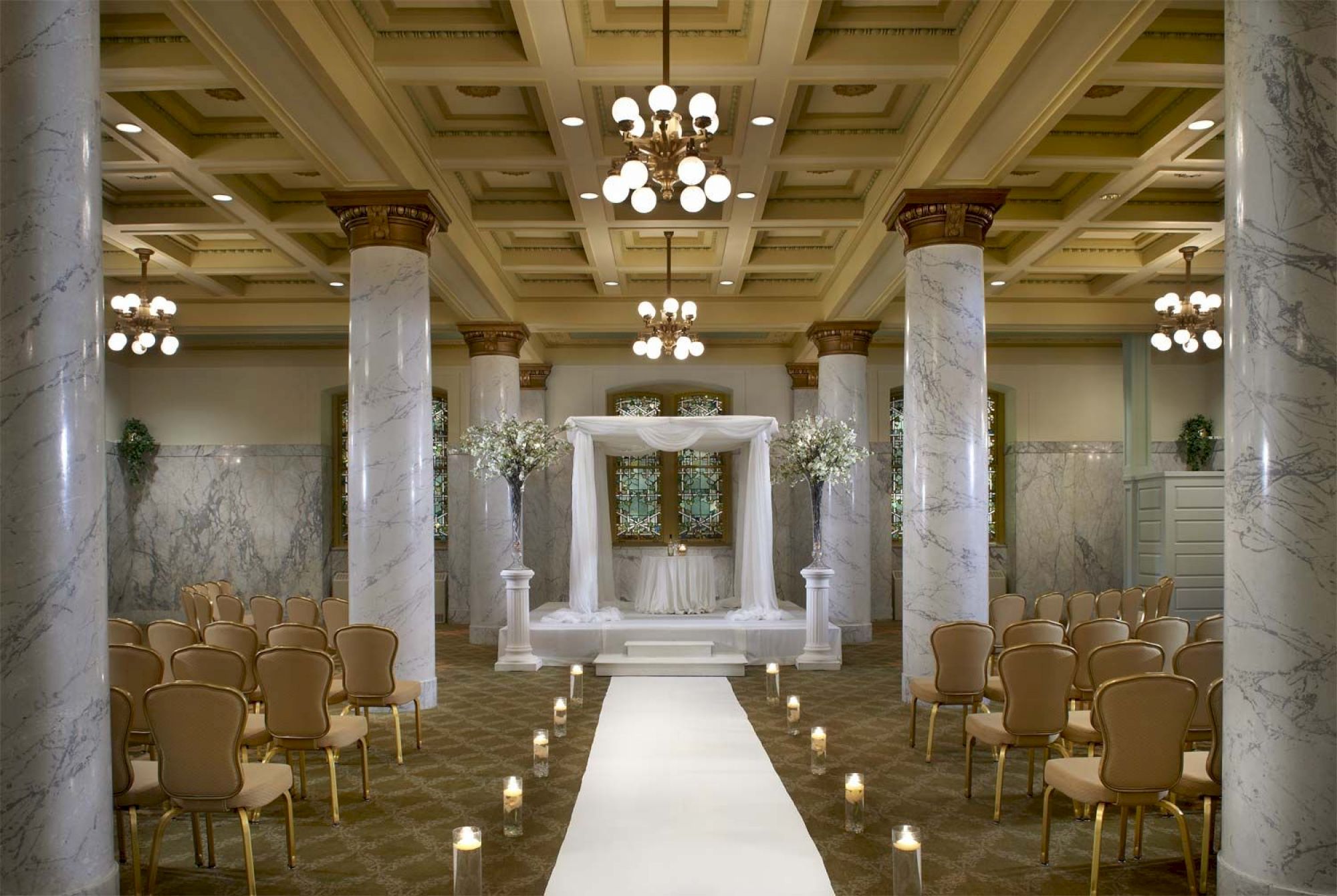 The image shows a decorated indoor wedding venue with a central aisle, chairs on both sides, a canopy at the front, marble columns, and chandeliers.