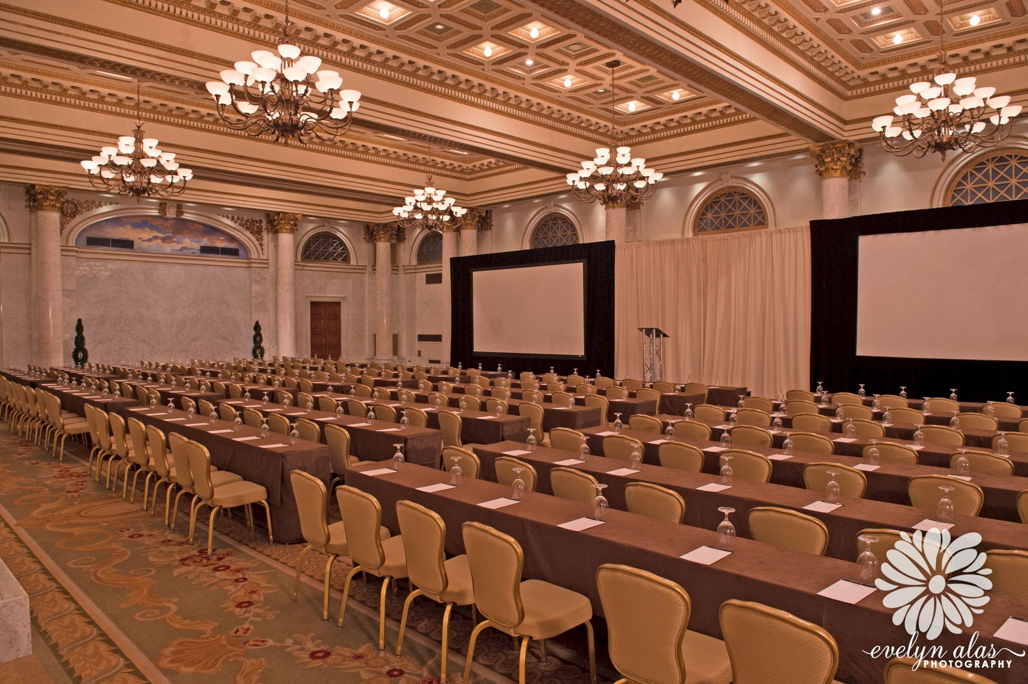 An elegantly decorated conference room with chandeliers, rows of chairs and tables, and two large projector screens. 