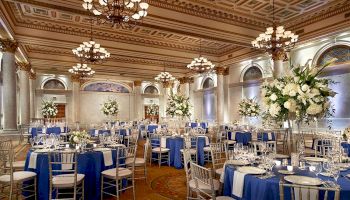 An elegant banquet hall with ornate chandeliers, blue and white table settings, tall floral centerpieces, and a luxurious, detailed ceiling.