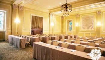 A decorated conference room with rows of chairs, tables, notepads, and pens, with chandeliers and wall lights illuminating the space.