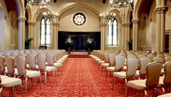 An elegant room with arched windows, chandeliers, red carpet, and rows of white chairs facing a stage, possibly set for a ceremony or event.