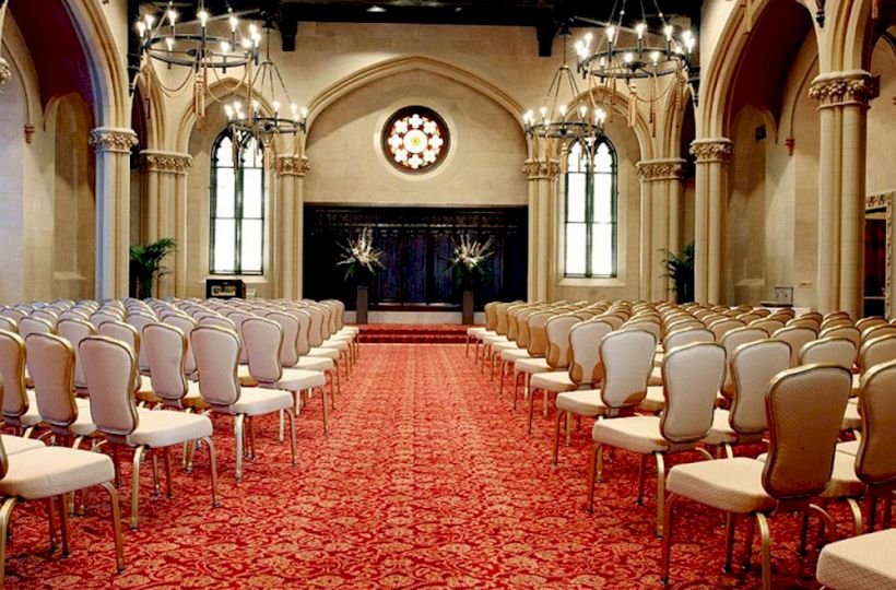 An elegant room with arched windows, chandeliers, red carpet, and rows of white chairs facing a stage, possibly set for a ceremony or event.