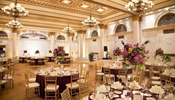 An elegantly decorated banquet hall with round tables, golden chairs, chandeliers, and large flower centerpieces, ready for a formal event or wedding.