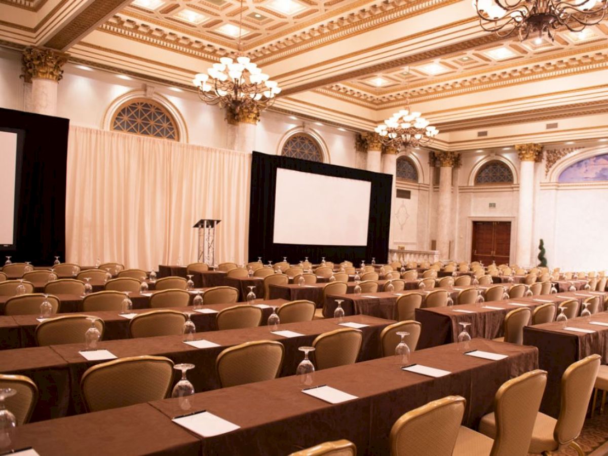 An elegant conference room is set up with rows of tables and chairs, notepads, and glasses, featuring two large projection screens and chandeliers.