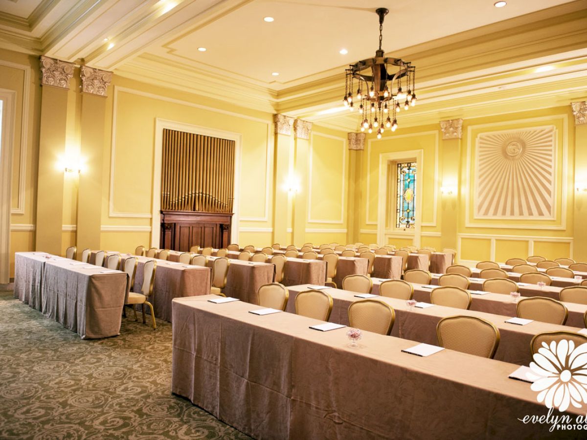 A well-lit conference room with rows of tables and chairs, intricate decor, and stained-glass windows at the back.