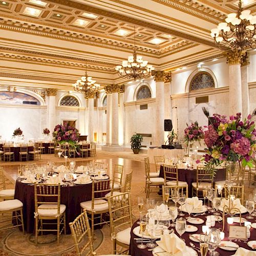 A grand, elegant ballroom set for a formal event, with round tables dressed with purple and white decor, gold chairs, and ornate chandeliers.