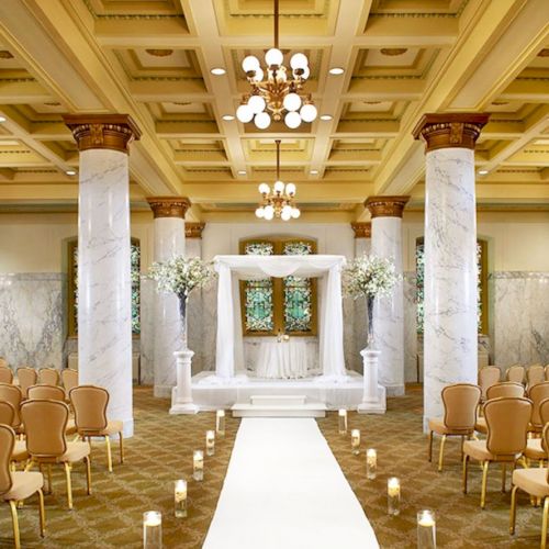 This is an elegant indoor wedding venue featuring marble columns, rows of chairs, a central aisle with candles, and a decorated altar at the front.