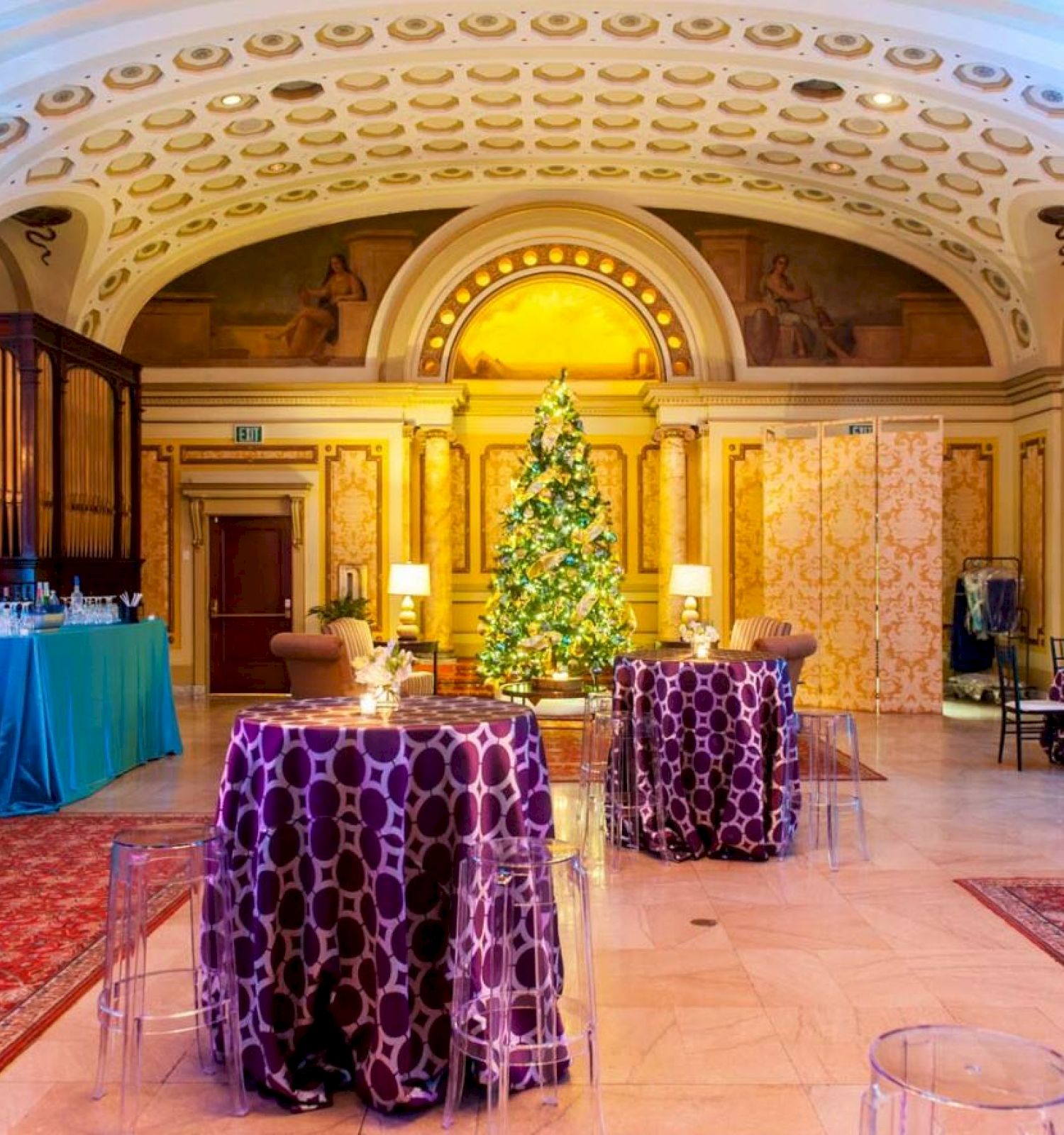 An elegant room featuring a decorated Christmas tree, tables with purple cloths, food stations, ornate ceiling, and classical architecture.