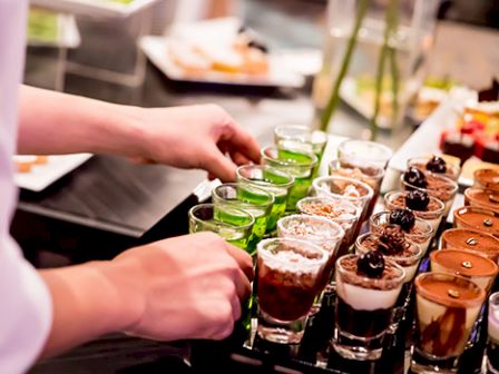 A person arranging a tray of assorted desserts in small glasses, including green jelly and chocolate mousse, in a buffet setting.