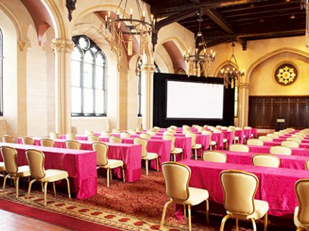 A conference room with long tables covered in red tablecloths, beige chairs, chandeliers, arched windows, and a large screen at the front.