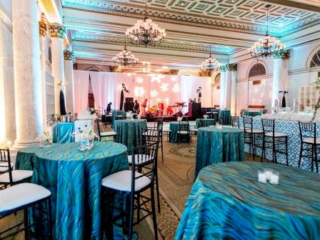 This image shows a decorated event space with round tables covered in turquoise cloth, chairs, chandeliers, and a stage set up with lights and instruments.